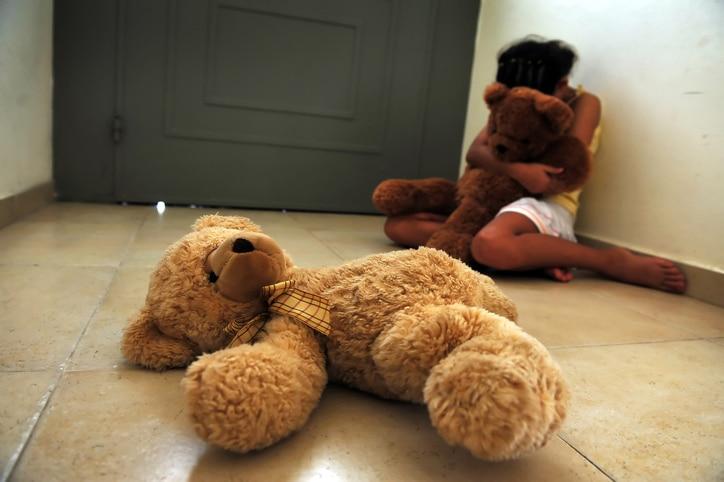 A young girl who is a victim of domestic violence sits on the floor next to the front door and gets comfort from her teddy bears