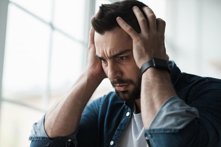 Sad and worried man sit at home couch feeling negative emotions, depression and problems. Upset excited young attractive bearded guy freaking out hold head with hands, on window background, close up