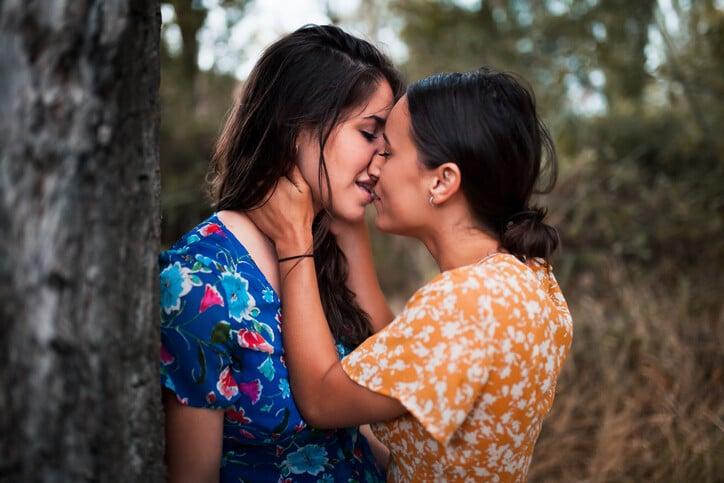 Two women kissing in the woods with dresses touching her face
