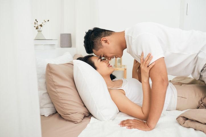 Side view of excited man is lying over woman in bed. They are looking at each other eyes and laughing