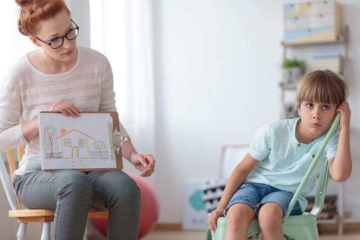 Young supported adoption counselor showing a drawing of house to sad pensive little boy waiting for new loving family